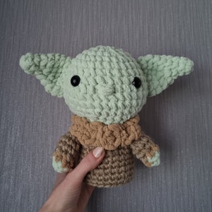May include: A crocheted doll of a character with large ears, black eyes, and a tan and green color scheme. The doll is held by a person. The doll's head and ears are light green, and the body is tan with green accents.