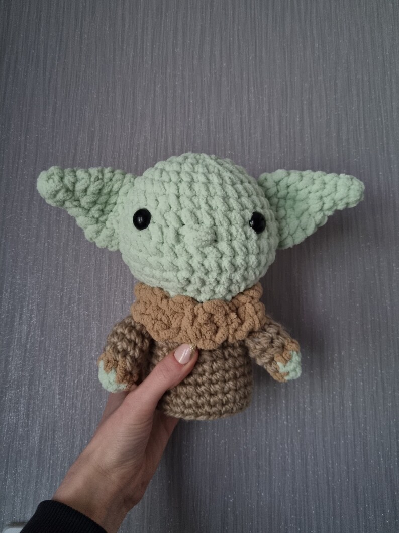 May include: A crocheted doll of a character with large ears, black eyes, and a tan and green color scheme. The doll is held by a person. The doll's head and ears are light green, and the body is tan with green accents.
