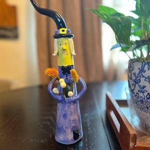 May include: A tall, slender ceramic figurine of a witch with a lime green face, wearing a tall black pointed hat with a yellow band. The figure has a purple dress, a black bow tie, and holds small yellow and white dried flowers in its arms.