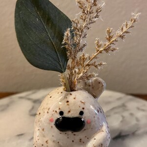 May include: A small, round, speckled ceramic figurine with a glossy finish, featuring two black eyes, a wide black mouth, and tiny pink cheeks. A single dark green leaf and a sprig of dried, textured beige grass are inserted into the top opening.