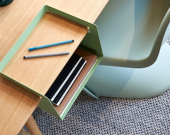 May include: A sage green metal desk organizer with a light wood grain surface. The organizer features a flat top tray holding two pens and a lower compartment containing two black spiral-bound notebooks and a brown notebook, attached to a desk.