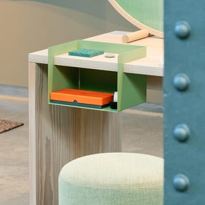 Könnte beinhalten: Ein hellgrünes Schreibtischregal aus Metall, das an einem hellen Holztisch befestigt ist. Das Regal enthält eine leuchtend orangefarbene rechteckige Schachtel und ein kleines schwarzes Objekt. Auf dem Tisch liegen ein kleiner grüner Fliesenrest und eine graue Scheibe.