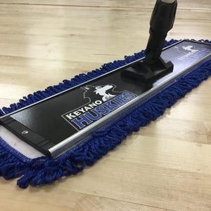 May include: A rectangular dust mop featuring a black base plate with the text KEYANO HUSKIES and a white husky logo. The mop head is surrounded by a thick, blue looped yarn fringe and includes a black plastic swivel handle attachment.