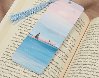 May include: A bookmark featuring a serene beach scene with a person on a surfboard, set against a gradient sky. The bookmark has a light blue tassel attached, and is placed in an open book.