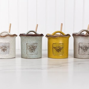 May include: Four ceramic honey pots with lids and wooden dippers. Each pot features a ruffled rim, a speckled finish, and a square embossed bee emblem. The pots are colored in white, light blue, mustard yellow, and light gray, arranged in a row.