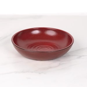 May include: A deep red ceramic bowl with a slightly rippled surface. The bowl is empty and sitting on a white marble surface.