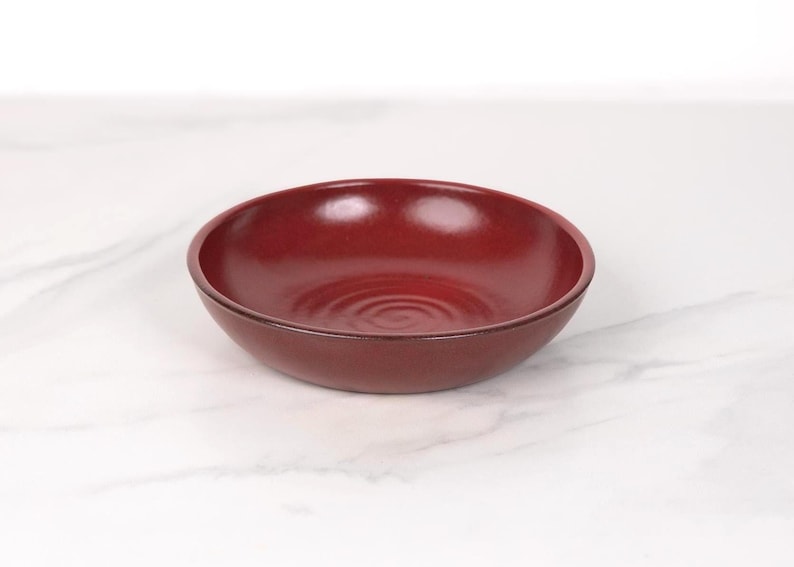May include: A deep red ceramic bowl with a slightly rippled surface. The bowl is empty and sitting on a white marble surface.