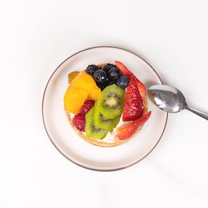 May include: A round fruit tart topped with sliced kiwi, strawberries, blueberries, raspberries, and orange segments on a white plate with a silver spoon. The pastry features a creamy white filling and a golden brown crust.