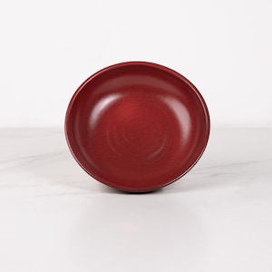 May include: A deep red ceramic bowl with a slightly textured surface. The bowl is empty and sitting on a white surface.