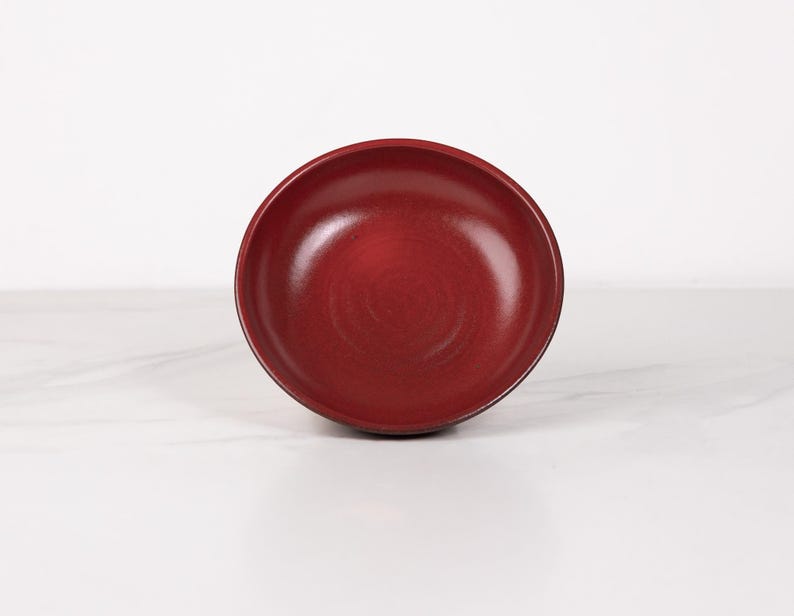 May include: A deep red ceramic bowl with a slightly textured surface. The bowl is empty and sitting on a white surface.