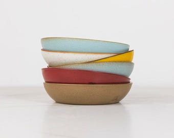 May include: A stack of five ceramic bowls in various colors including light blue, white, yellow, red, and brown. Each bowl features a speckled texture and a smooth, rounded rim. The bowls are arranged vertically, creating a colorful display of kitchenware.