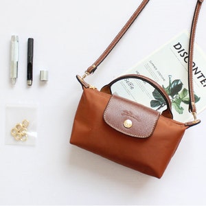 May include: A brown leather and nylon handbag with a long strap and a top handle. The bag has a gold-coloured zipper and hardware. The bag is sitting on a white surface with a magazine that says "DISCONTENT" on it.