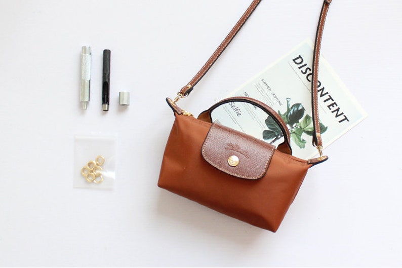 May include: A brown leather and nylon handbag with a long strap and a top handle. The bag has a gold-coloured zipper and hardware. The bag is sitting on a white surface with a magazine that says "DISCONTENT" on it.