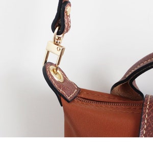 May include: Close-up of a brown leather handbag with a gold metal clasp and a zip. The bag has a brown nylon lining and a black strap.