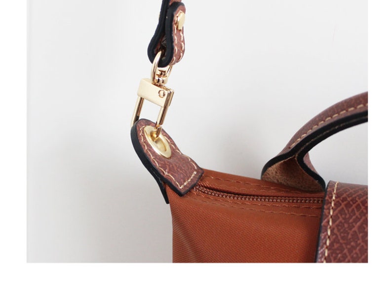 May include: Close-up of a brown leather handbag with a gold metal clasp and a zip. The bag has a brown nylon lining and a black strap.