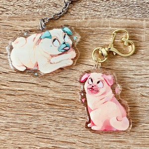 May include: Two clear acrylic keychains featuring cartoon pug illustrations. One pug is laying down with a light blue collar and the other is sitting up with a pink collar. Both pugs have glitter accents.