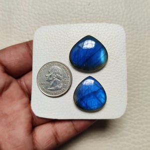 Puede incluir: Dos piedras preciosas de labradorita pulidas en forma de lágrima, con iridiscencia azul y dorada. Una moneda de cuarto de dólar estadounidense se encuentra junto a las piedras preciosas para comparar el tamaño. Las piedras preciosas se muestran sobre una superficie blanca.