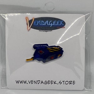 May include: A blue and yellow Ghostbusters proton pack enamel pin with a red light on the side. The pin is on a white card with the text "Vendageek" and "www.vendageek.store".