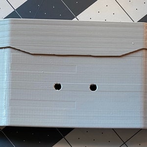 May include: A light gray, rectangular 3D-printed object with a series of holes. The object has a notched top and appears to be a housing or enclosure. The surface has visible layer lines from the printing process.