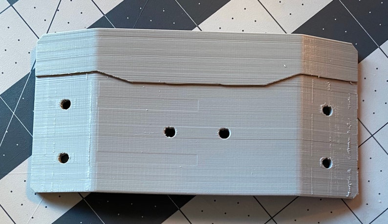 May include: A light gray, rectangular 3D-printed object with a series of holes. The object has a notched top and appears to be a housing or enclosure. The surface has visible layer lines from the printing process.