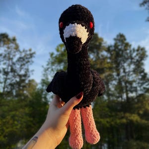 May include: A handmade black crochet bird with a white patch on its face and red eyes. The bird has pink legs and is held against a backdrop of trees and a blue sky. The plush toy is soft and detailed.