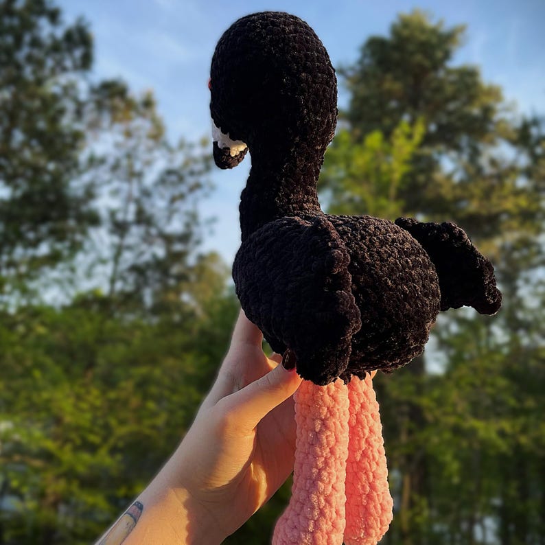 May include: A handmade black crochet ostrich with a white beak and red accents. The ostrich has pink legs and is held up against a blurred green background. The plush toy is made of soft yarn.