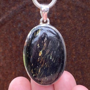 May include: Oval pendant with a polished black stone, featuring streaks of gold and silver, set in a silver bezel. The pendant hangs from a silver chain. The gemstone is approximately 1.5 inches long.