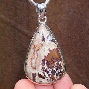 May include: A teardrop-shaped pendant with a silver bezel and bail. The stone within features a mix of white, brown, and burgundy colours. The pendant hangs from a silver chain, creating a striking contrast.