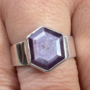 Può includere: Primo piano di un anello d'argento con una grande gemma esagonale rosso-viola. La fascia dell'anello è larga e liscia, e la gemma ha un motivo a stella. L'anello è su un dito.