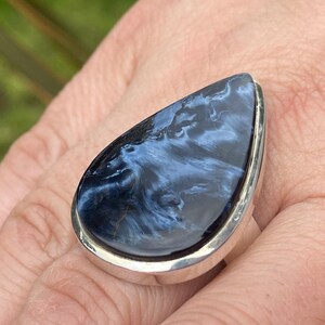 May include: A large, teardrop-shaped ring featuring a polished blue gemstone with swirling patterns. The stone is set in a silver band, creating a striking contrast. The ring is displayed on a hand, showcasing its size and design.