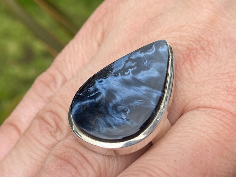 May include: A large, teardrop-shaped ring featuring a polished blue gemstone with swirling patterns. The stone is set in a silver band, creating a striking contrast. The ring is displayed on a hand, showcasing its size and design.