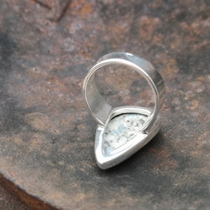 May include: A silver ring with a white and gray stone set in a teardrop shape. The ring is shown from the back, with the band visible.