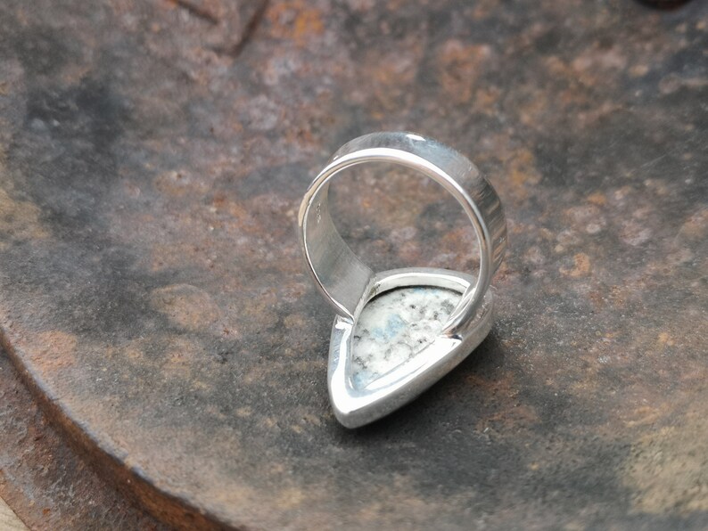 May include: A silver ring with a white and gray stone set in a teardrop shape. The ring is shown from the back, with the band visible.