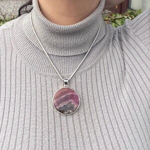 May include: A silver chain necklace with a round pendant featuring a pink and purple gemstone. The pendant has a layered, striated pattern.