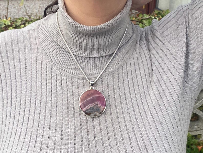 May include: A silver chain necklace with a round pendant featuring a pink and purple gemstone. The pendant has a layered, striated pattern.