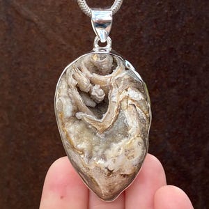May include: A natural stone pendant with a silver-coloured bail and chain. The pendant has a teardrop shape and features a mix of beige, grey, and white tones. The stone has a textured, organic appearance, with a polished silver border.
