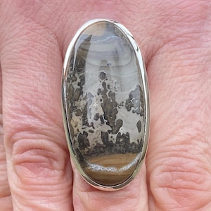 May include: Oval-shaped ring with a large, polished stone. The stone features a landscape-like pattern in shades of gray, brown, and white, set in a silver-colored metal band. The ring is worn on a finger.