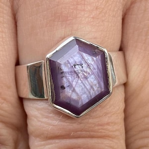 May include: A silver ring featuring a large, faceted, hexagonal purple gemstone. The gemstone has a star-like pattern within it. The ring's band is wide and made of polished silver, creating a bold and elegant look.
