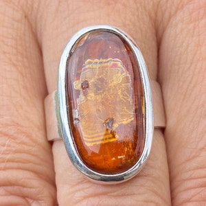 May include: A silver ring with an oval-shaped amber stone. The stone has a golden hue with iridescent streaks.