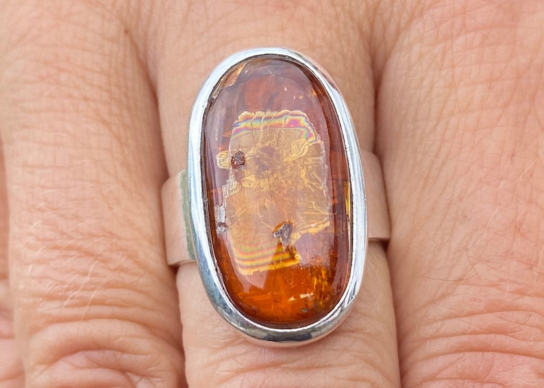 May include: A silver ring with an oval-shaped amber stone. The stone has a golden hue with iridescent streaks.