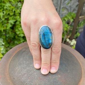 May include: A silver ring with a large oval blue labradorite gemstone. The ring is on a finger and the stone has a iridescent sheen.