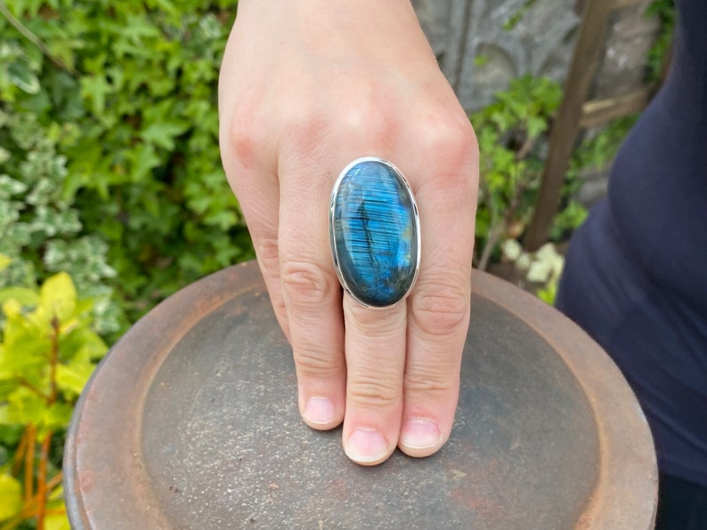 May include: A silver ring with a large oval blue labradorite gemstone. The ring is on a finger and the stone has a iridescent sheen.