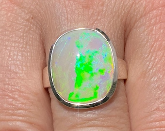 May include: A silver ring featuring a large, oval opal gemstone. The opal displays vibrant green, yellow, and blue flashes against a milky white base. The ring has a simple, polished band.
