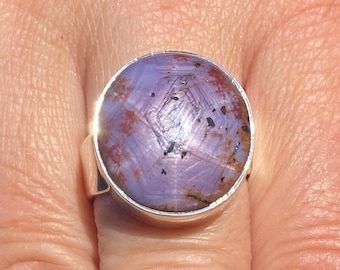 May include: A silver ring featuring a large, round, cabochon-cut stone with a lavender and purple hue. The stone displays a natural star-like pattern across its surface, accented by dark brown and black inclusions throughout the translucent material.