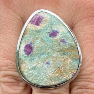 以下が含まれることがあります： 緑と金の宝石が涙の形にセットされたシルバーリング。宝石にはピンクのルビーのインクルージョンがあります。