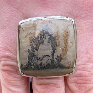 May include: A square, silver-toned ring featuring a large, polished stone with a unique pattern. The stone displays a blend of beige, brown, and black hues, creating a natural, artistic design. The ring's setting is a simple, polished silver.