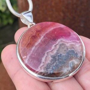 May include: A silver pendant with a round, pink and gray rhodochrosite stone. The stone has a swirling pattern of pink, white, and gray.