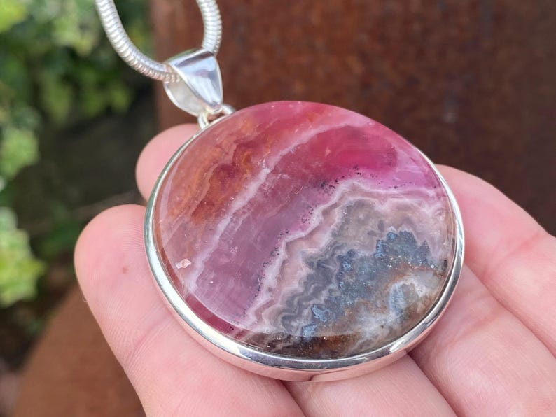May include: A silver pendant with a round, pink and gray rhodochrosite stone. The stone has a swirling pattern of pink, white, and gray.