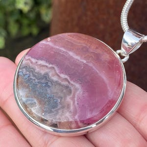 May include: A round pink and gray gemstone pendant with a silver setting and chain. The pendant is approximately 2 inches in diameter.