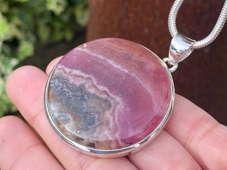 May include: A round pink and gray gemstone pendant with a silver setting and chain. The pendant is approximately 2 inches in diameter.